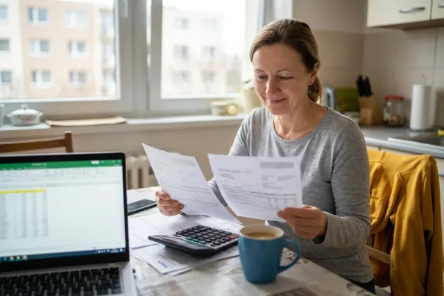 Žena u kuchyňského stolu porovnává dvě vyúčtování elektřiny a počítá na kalkulačce vedle notebooku.