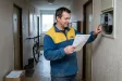 Muž u elektroměru v chodbě panelového domu porovnává vytištěné vyúčtování elektřiny.