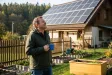 Muž na zahradě rodinného domu se dívá na fotovoltaické panely na střeše a drží hrnek s kávou.