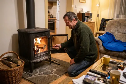 Muž přikládá poleno do hořících krbových kamen v obývacím pokoji za zimního večera.