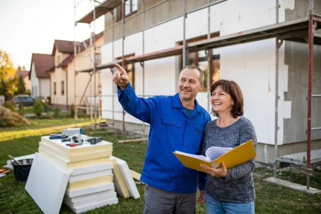 Pár se usmívá před rodinným domem s lešením a izolačními deskami na fasádě, žena drží složku s papíry.