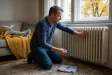 Muž zkouší teplotu radiátoru pod oknem a chystá se odvzdušňovacím klíčem upravit ventil.