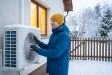 Muž v zimní bundě kontroluje zasněženou venkovní jednotku tepelného čerpadla u domu, v okně svítí teplé světlo.